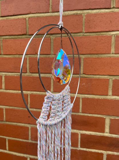 Handmade macrame Suncatcher made with pastel coloured recycled yarn, on two metal hoops, with a large hanging crystal in the centre, hanging outside in front of a brick wall.