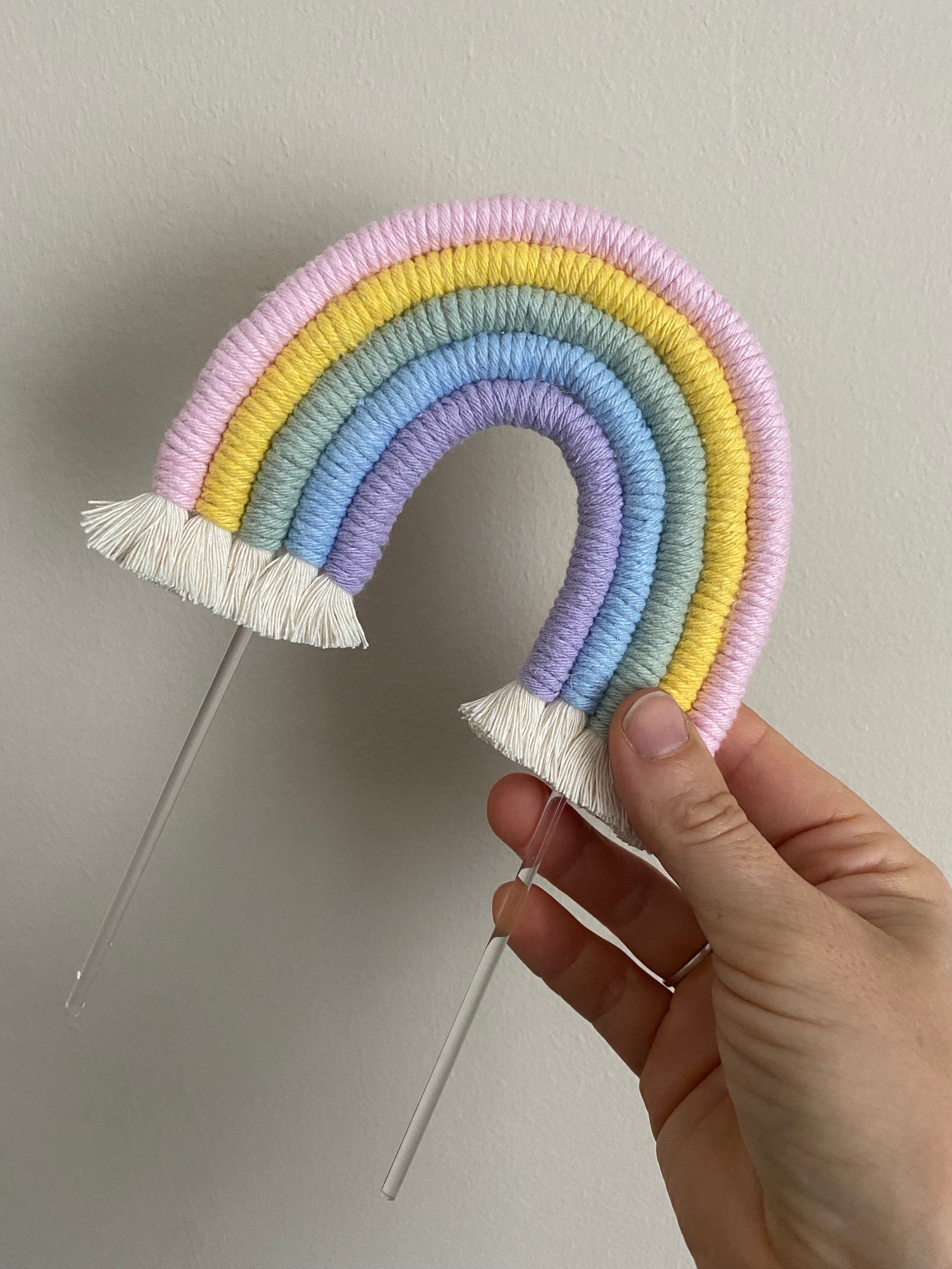 A hand holding a pastel rainbow macramé cake topper with long clear acrylic sticks, in front of a beige painted wall. 