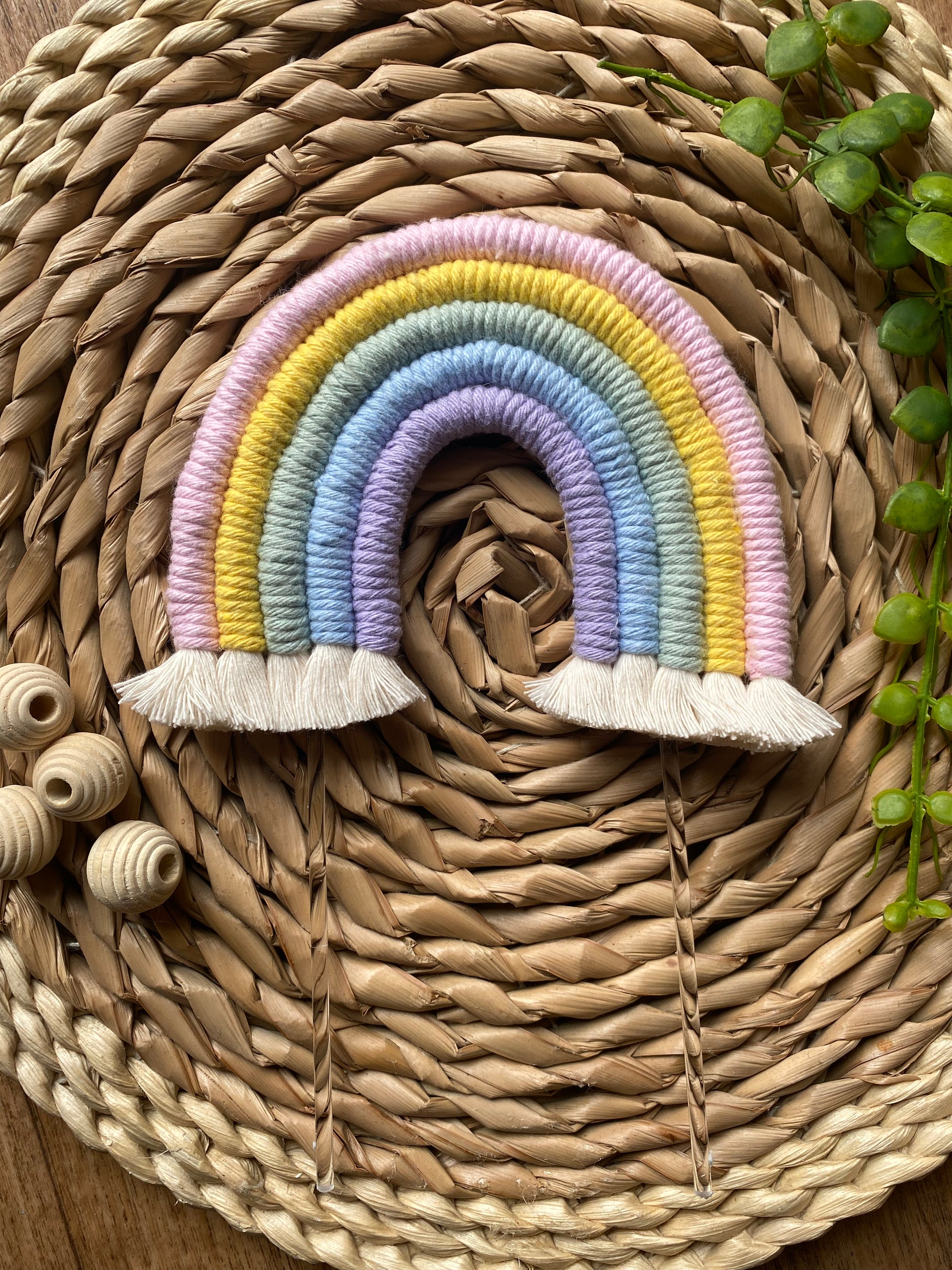 Pastel rainbow macramé cake topper with long clear acrylic sticks, laid on a round wicker mat with wooden beads nearby for decoration. 