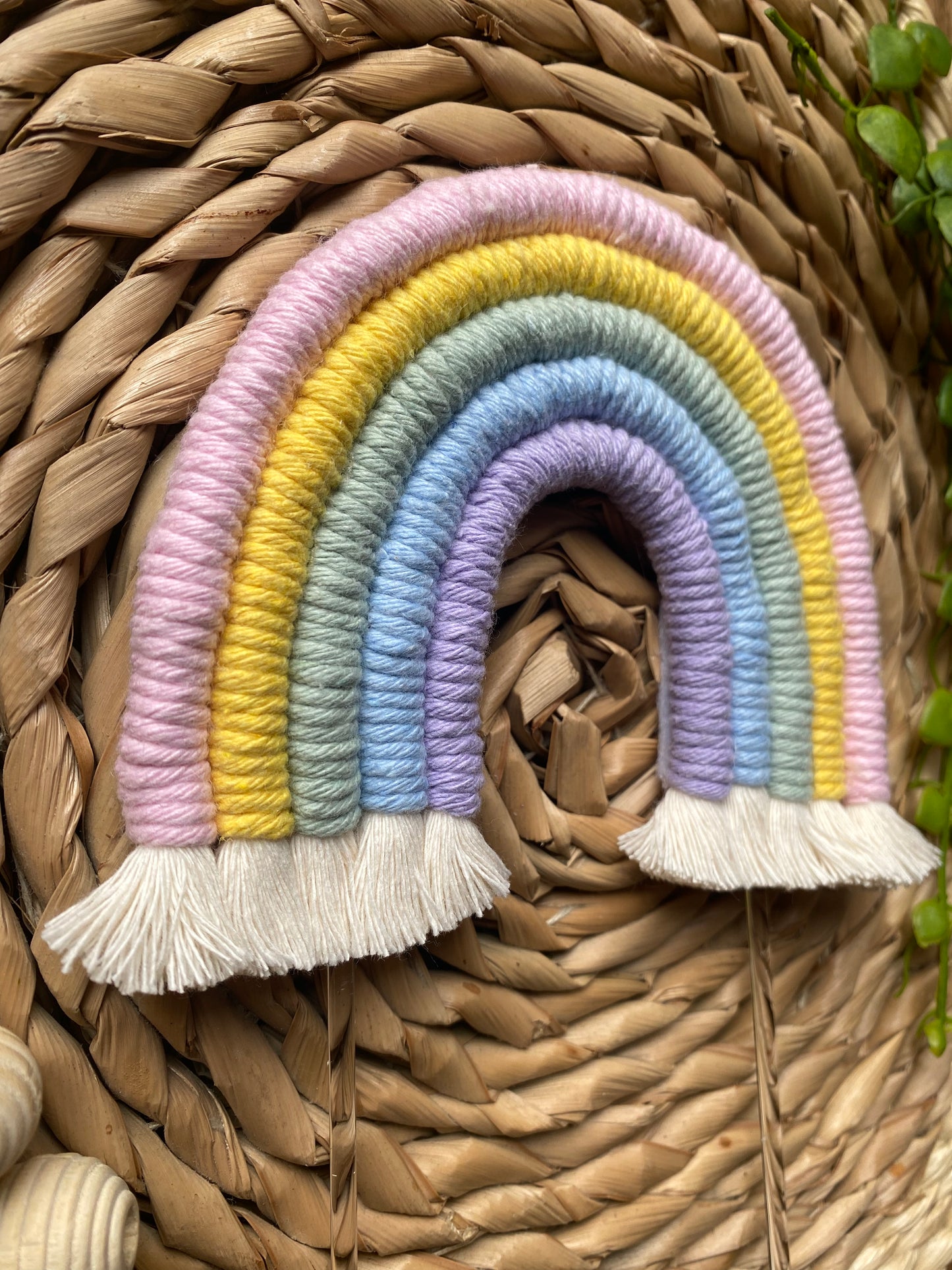 Close up of a pastel rainbow macramé cake topper with long clear acrylic sticks, laid on a round wicker mat with wooden beads nearby for decoration. 