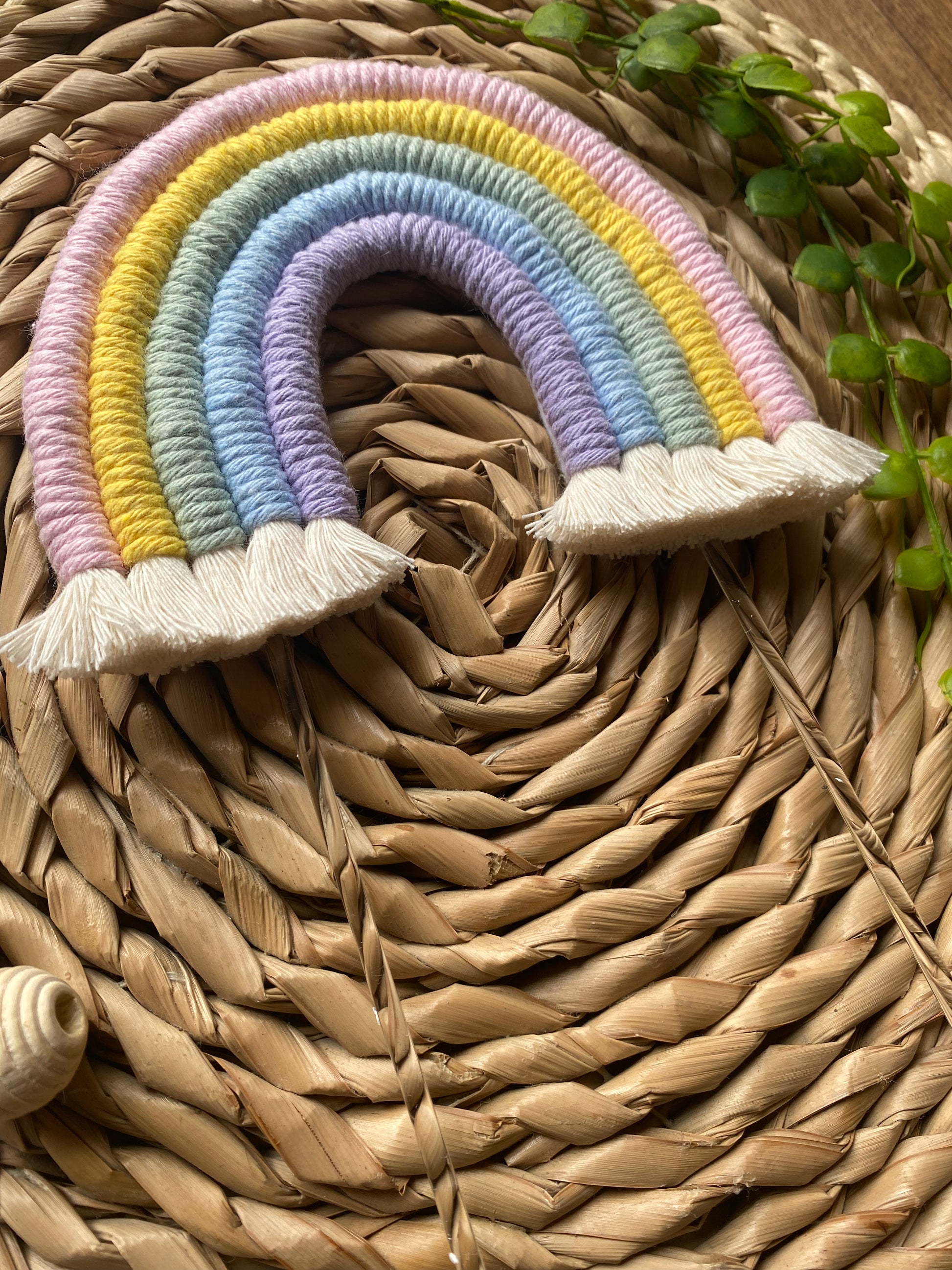 Pastel rainbow macramé cake topper with long clear acrylic sticks, laid on a round wicker mat.