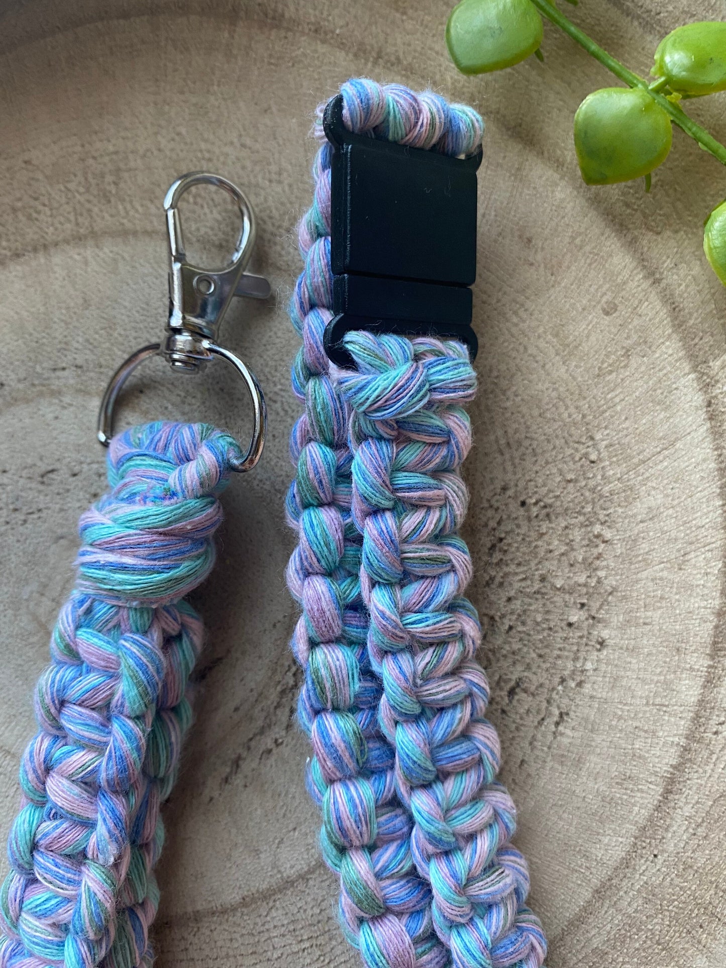 A handmade, colorful woven Eco friendly lanyard badge holder - 34” with a metal swivel hook clasp on one end and a black plastic buckle on the other. This eco-friendly lanyard by Macra-Made-With-Love features a braided pattern in pastel blue, green, and pink threads. It is placed on a wooden surface with some green plant decoration.