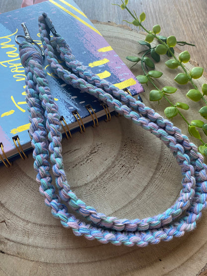 A colorful, handmade lanyard is displayed on a wooden surface, draped across a notebook with a patterned cover. An Eco friendly lanyard badge holder - 34” by Macra-Made-With-Love lies next to the notebook, adding a touch of nature to the composition with its green leafy plant.