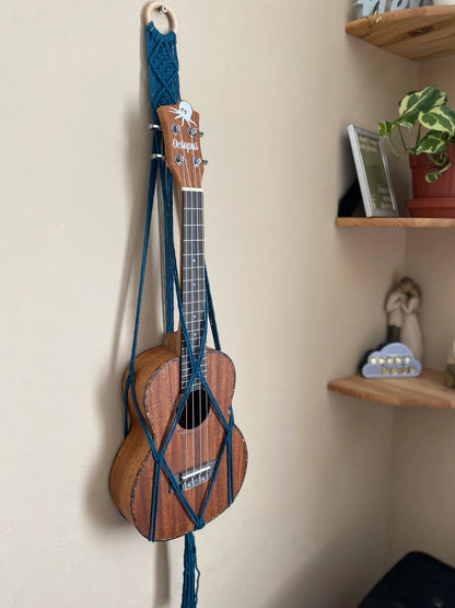 A dark brown ukulele sits inside of a handmade macrame wall hanging ukulele hanger, which is made with petrol blue recycled cotton, and a repurposed wooden hanging ring. 