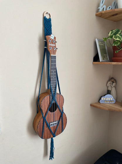 A dark brown ukulele sits inside of a handmade macrame wall hanging ukulele hanger, which is made with petrol blue recycled cotton, and a repurposed wooden hanging ring. A set of corner shelves is to the right of the image. 