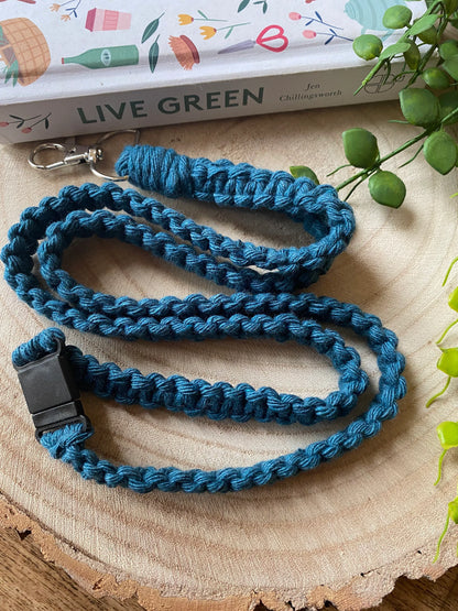 A handmade blue braided dog leash sits coiled on a wooden surface. There is a black plastic clip on one end and a metal clasp on the other. Above the leash, an Eco friendly lanyard badge holder - 34” by Macra-Made-With-Love lies next to "Live Green" by Jen Chillingsworth and a small plant, partially visible.