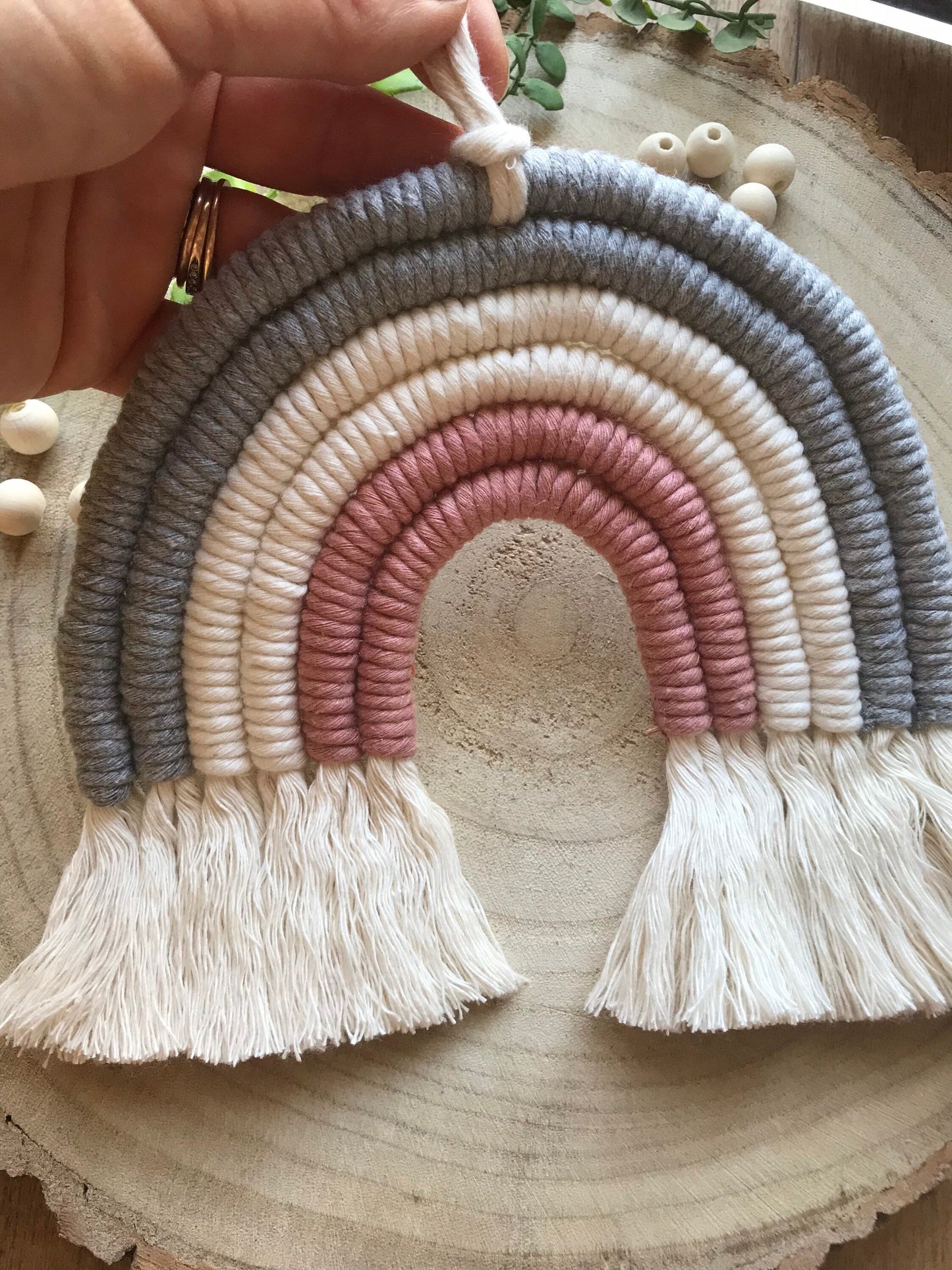 A hand holding a macrame rainbow made with 6 arches of grey, natural and pink yarn with combed out tassels at the bottom.