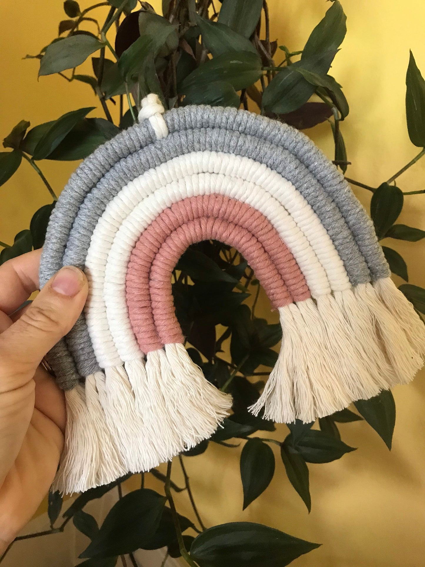 A hand holding a macrame rainbow made with 6 arches of grey, natural and pink yarn with combed out tassels at the bottom, in front of a trailing houseplant and a yellow wall. 