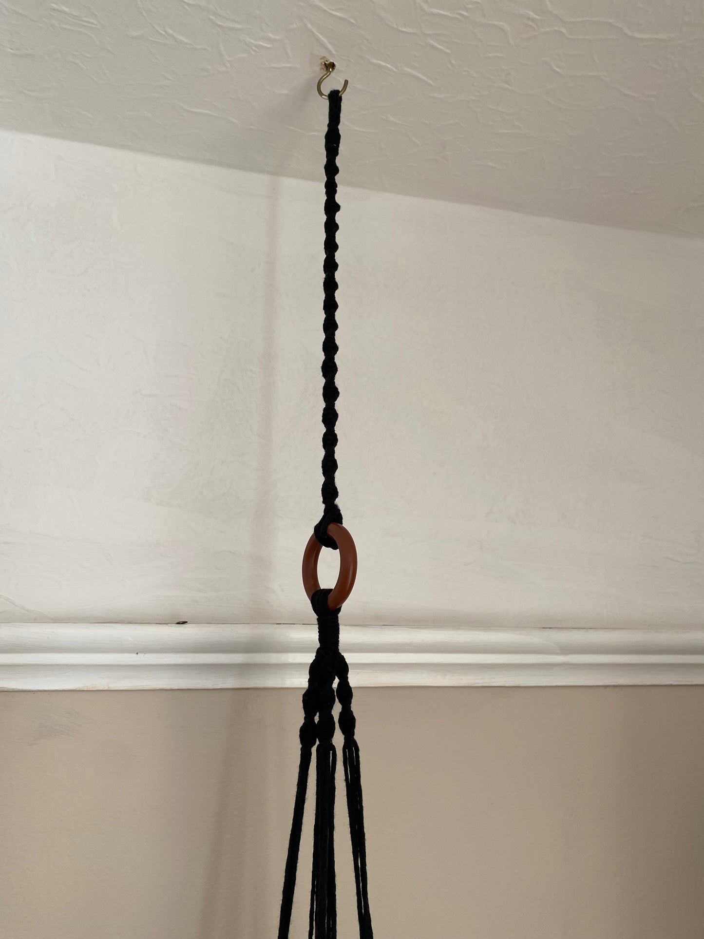 A macrame plant hanger extension made with black cotton yarn, is attached to the wooden ring of a macrame plant hanger, and suspended from a white ceiling on a metal hook. 