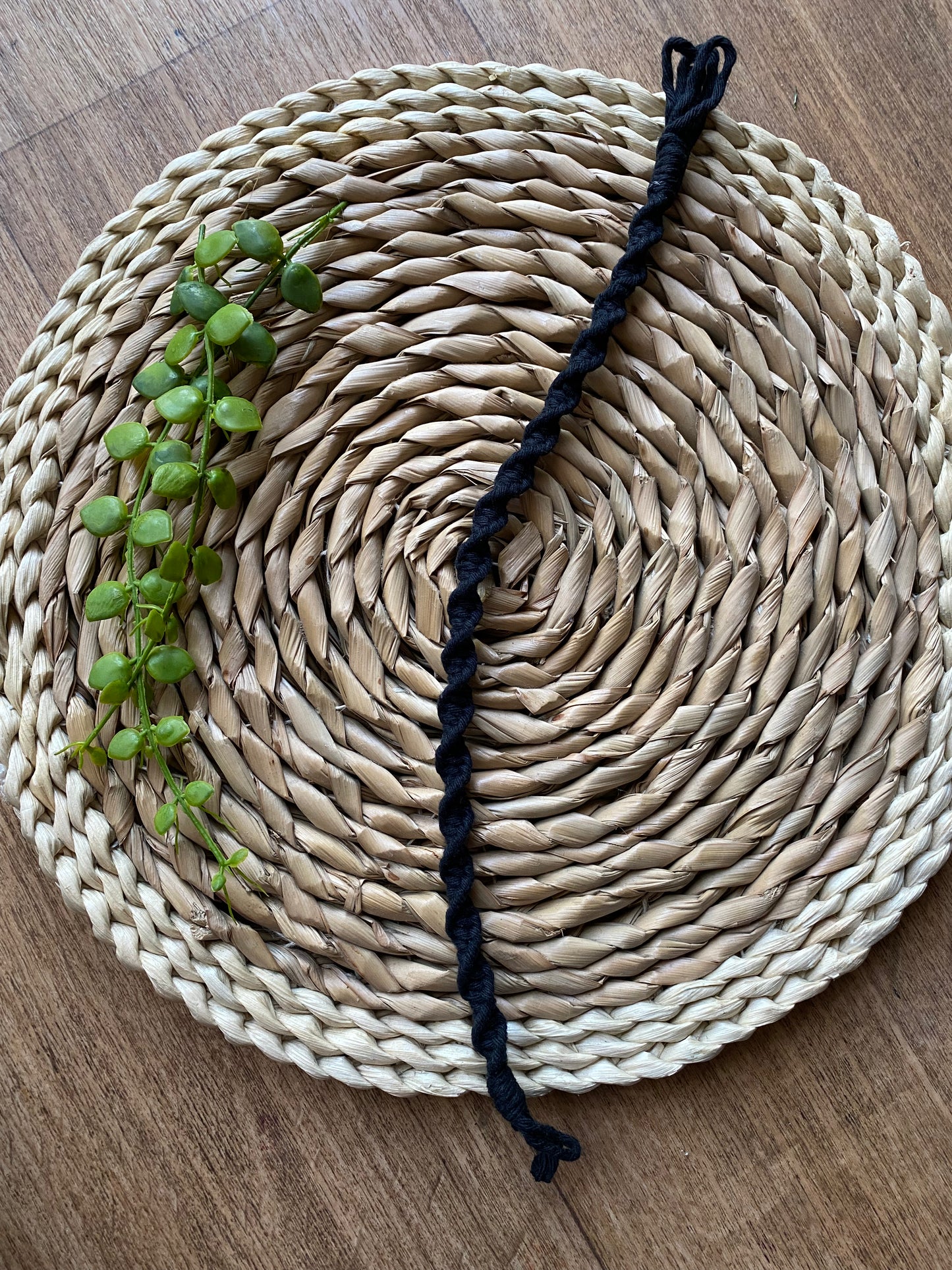 A long macrame plant hanger extension made with black cotton yarn, is laid out across a round wicker mat. 