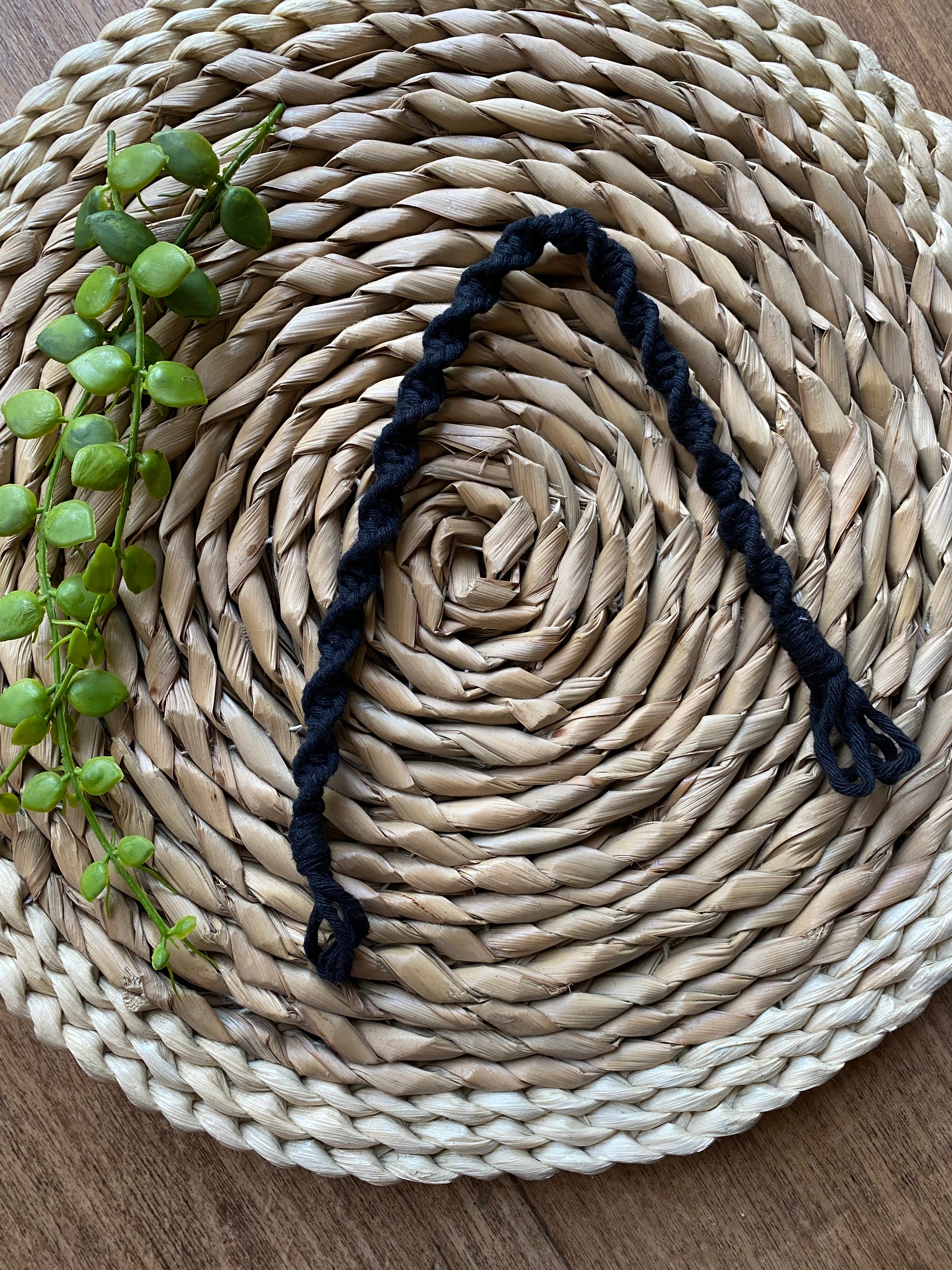 A macrame plant hanger extension made with black cotton yarn, is folded on a round wicker mat. 