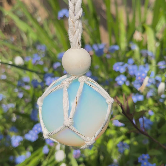 Video showing a handmade macrame opaline sphere hanging sun catcher, made with eco friendly natural recycled cotton yarn. The sphere is shown hanging outside in front of some small blue flowers, there is a plain wooden bead above the crystal.
