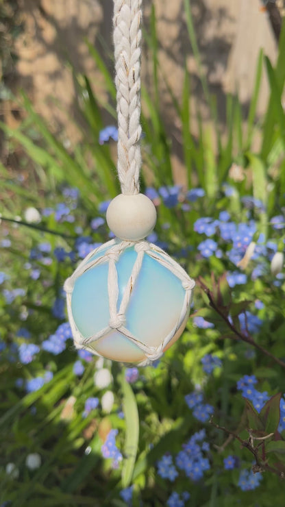 Video showing a handmade macrame opaline sphere hanging sun catcher, made with eco friendly natural recycled cotton yarn. The sphere is shown hanging outside in front of some small blue flowers, there is a plain wooden bead above the crystal.