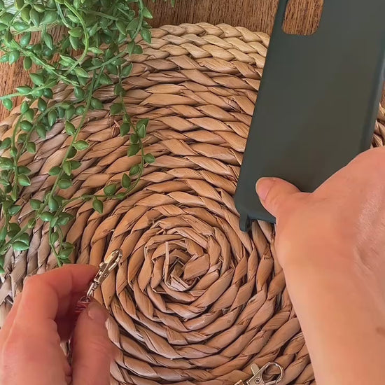 A video demonstrating how to use a handmade short macrame phone wristlet loop for phone case. Shown being attached to a green phone case on a wicker mat. Handmade with multicoloured recycled cotton yarn, with a small metal lobster clasp on each end. 