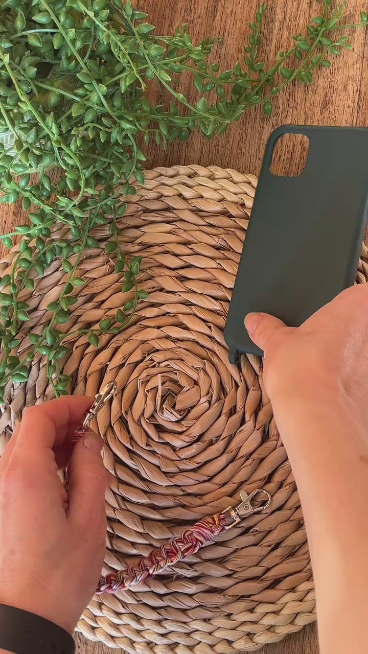 A video demonstrating how to use a handmade short macrame phone wristlet loop for phone case. Shown being attached to a green phone case on a wicker mat. Handmade with multicoloured recycled cotton yarn, with a small metal lobster clasp on each end. 