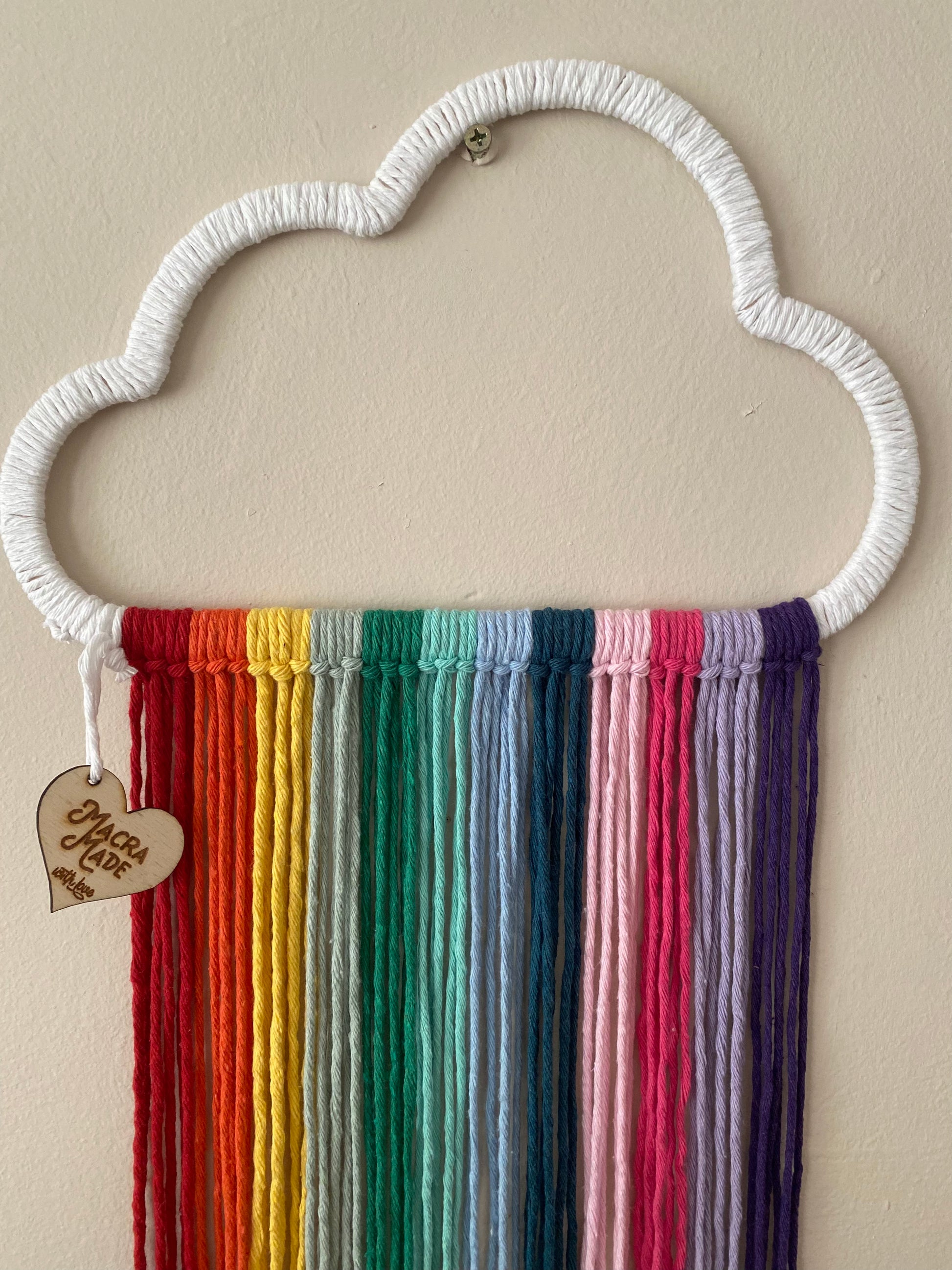 A cloud shaped wall hanging decoration, made with rainbow hanging tassels, displayed against a beige wall.