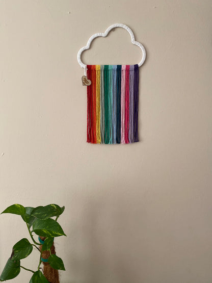 A cloud shaped wall hanging decoration, made with rainbow hanging tassels, displayed against a beige wall.