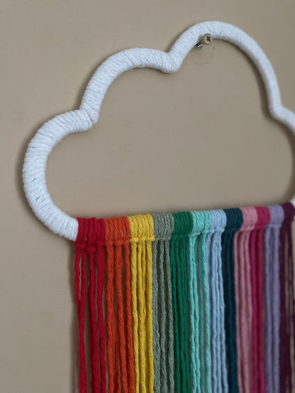 A close up view of a cloud shaped wall hanging decoration, made with rainbow hanging tassels, displayed against a beige wall.