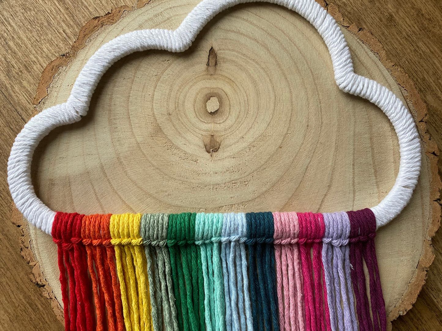 A cloud shaped wall hanging decoration, made with rainbow hanging tassels, displayed on a round wooden surface.