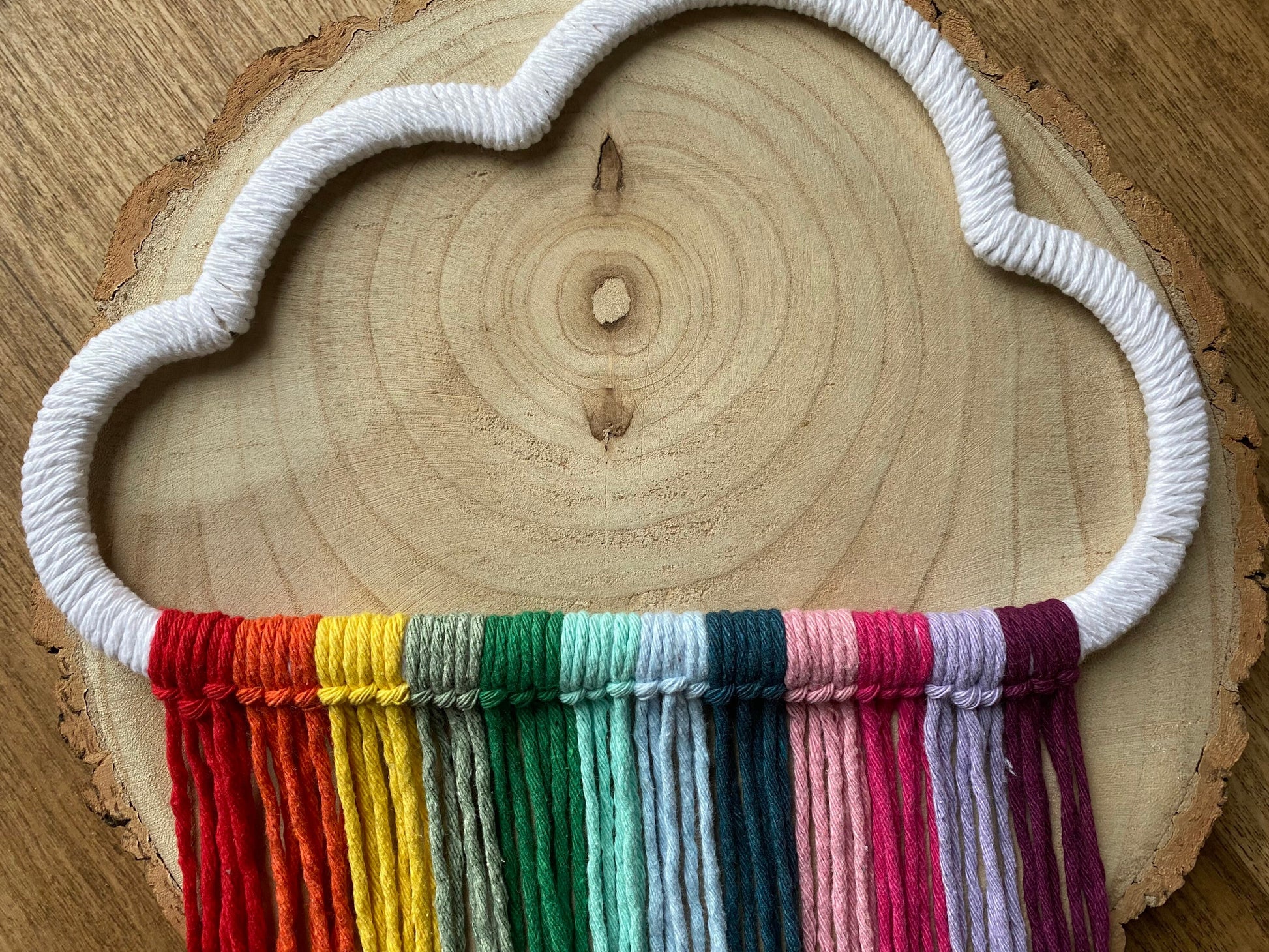 A cloud shaped wall hanging decoration, made with rainbow hanging tassels, displayed on a round wooden surface.