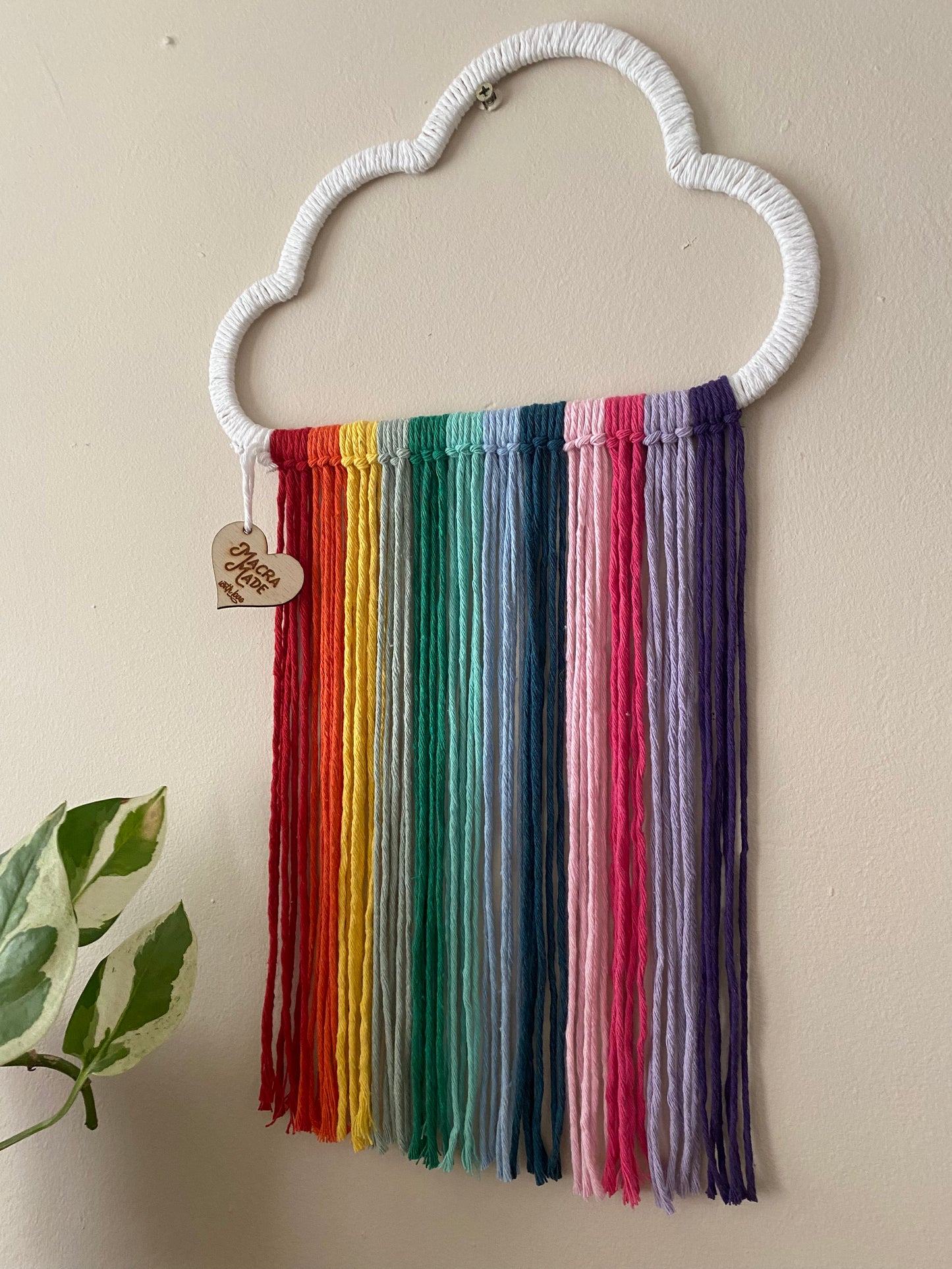 A close up view of a cloud shaped wall hanging decoration, made with rainbow hanging tassels, displayed against a beige wall.