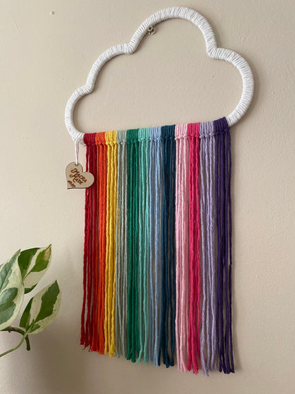 A close up view of a cloud shaped wall hanging decoration, made with rainbow hanging tassels, displayed against a beige wall.