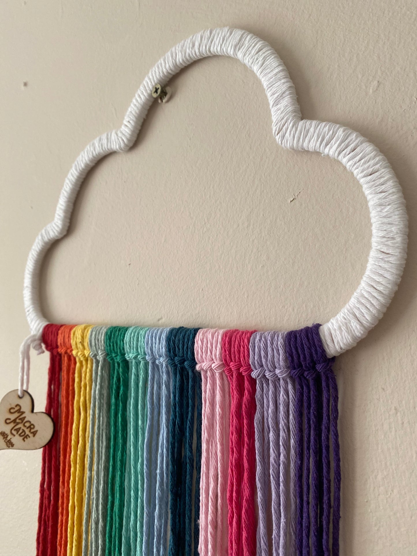 A close up view of a cloud shaped wall hanging decoration, made with rainbow hanging tassels, displayed against a beige wall.
