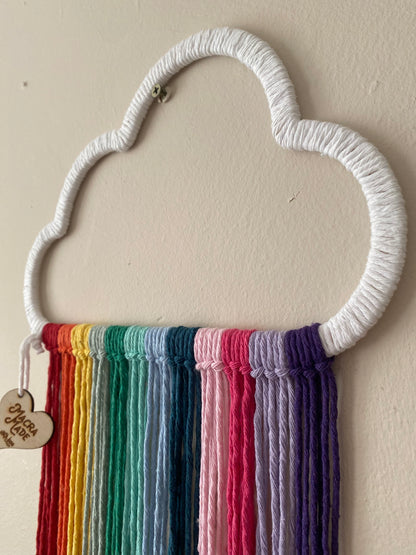 A close up view of a cloud shaped wall hanging decoration, made with rainbow hanging tassels, displayed against a beige wall.