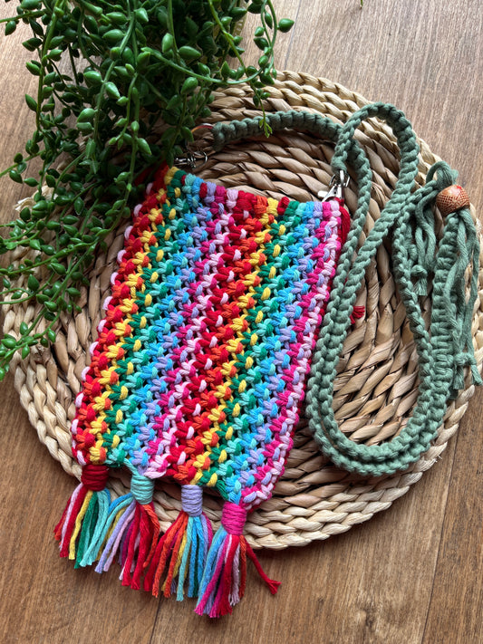 Colourful macrame crossbody bag with tassels at the bottom, and a green adjustable strap at the top, on a round wicker mat. 