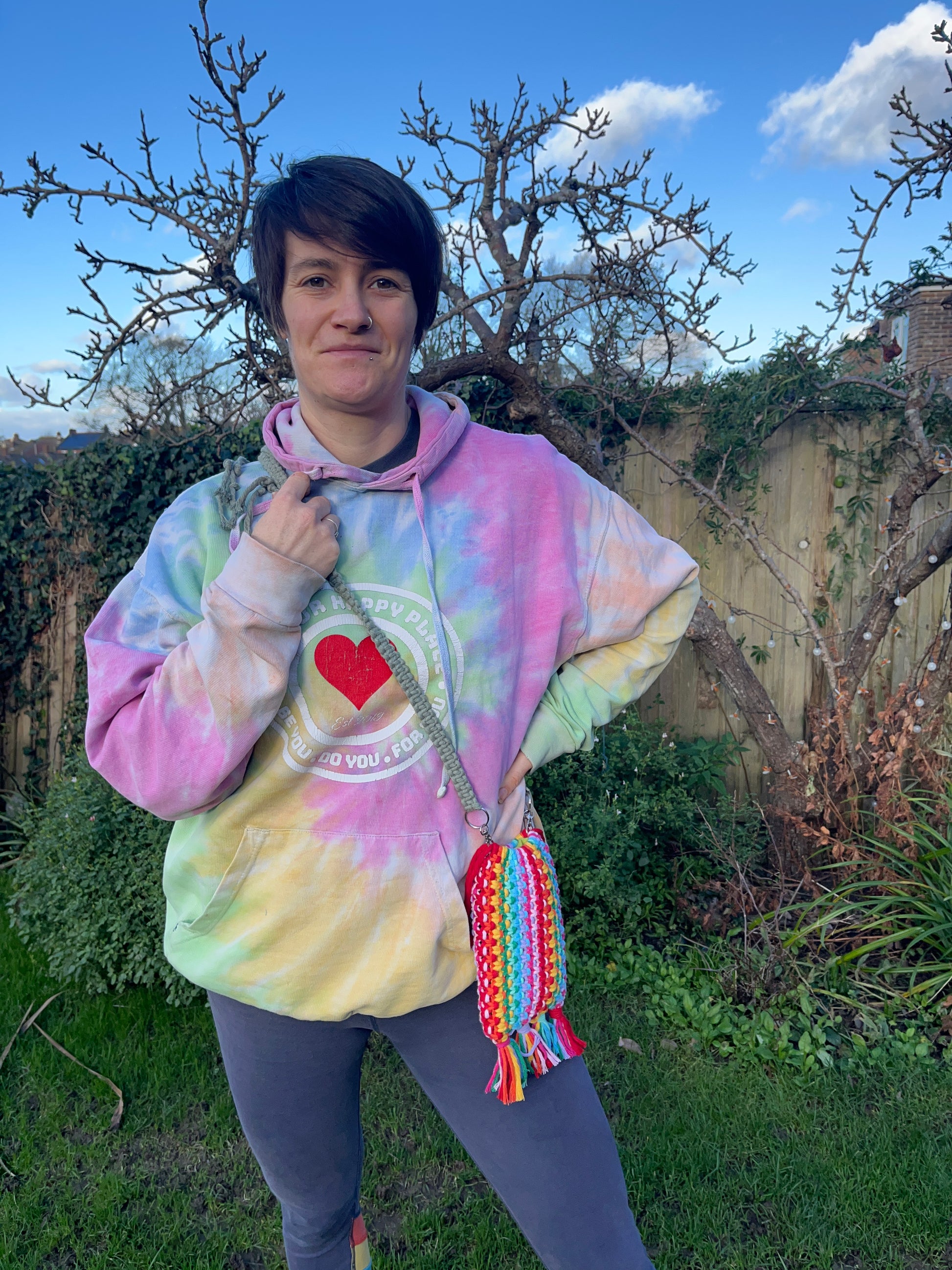 A woman standing outside wearing a rainbow hoodie, and a crossbody macrame bag, made in rainbow colours with recycled cotton yarn. 