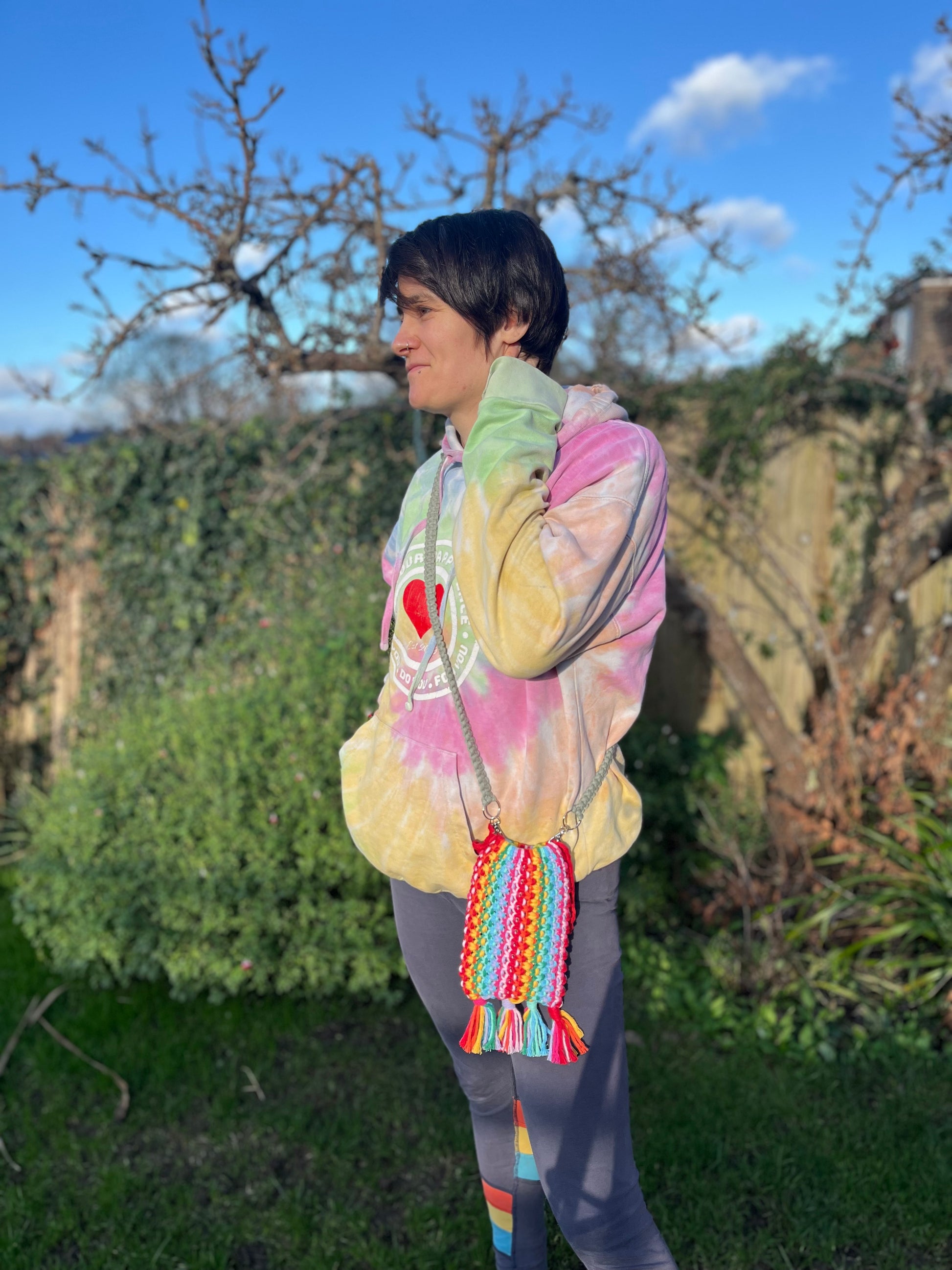 A woman stands outside wearing a rainbow hoodie, facing sideways on to the camera. She is wearing a crossbody bag, which is made with rainbow coloured yarn and a long green strap. 