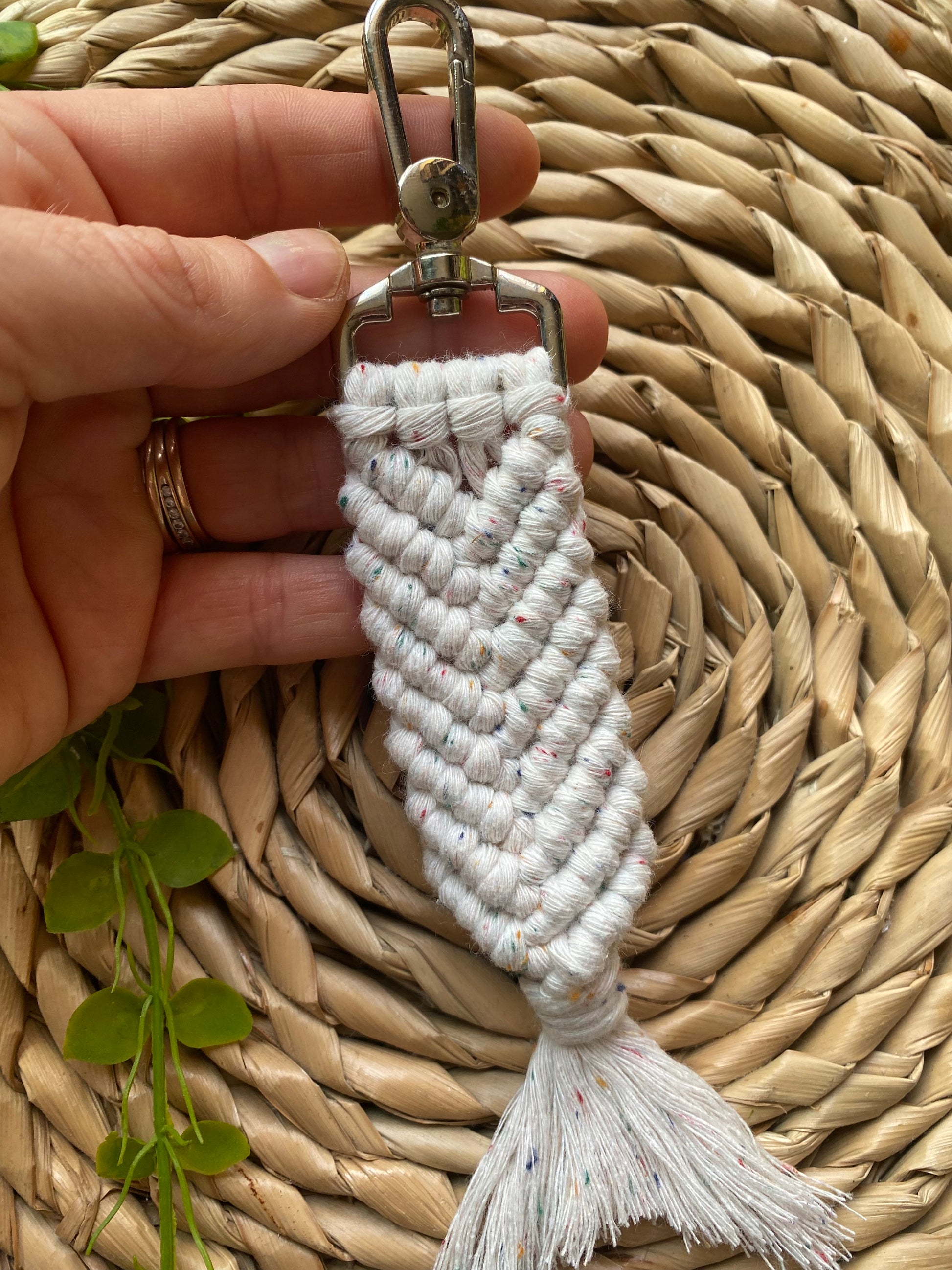 A hand holding a handmade macrame keyring, knotted using natural coloured recycled cotton, with rainbow flecks, in the shape of a mermaids tail, and finished with a metal lobster clasp. 