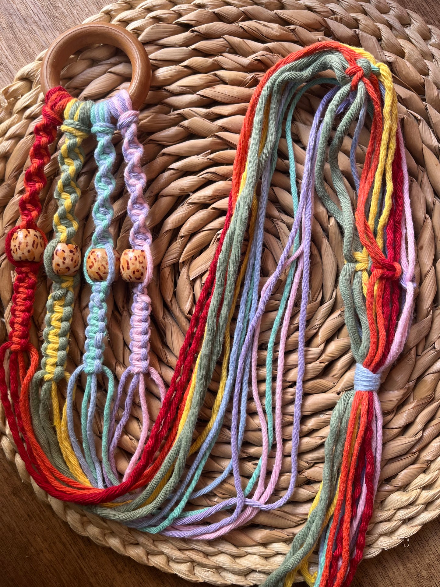 Handmade macramé plant hanger with patterned wooden beads and rainbow coloured yarns displayed on a woven mat.