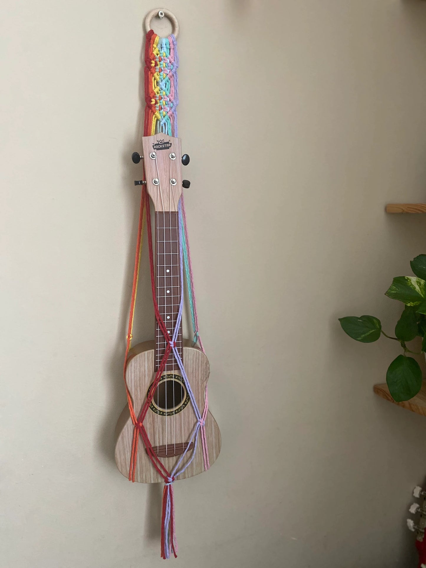 A light brown ukulele sits inside of a handmade macrame wall hanging ukulele hanger, which is made with rainbow colours of recycled cotton, and a repurposed wooden hanging ring. 