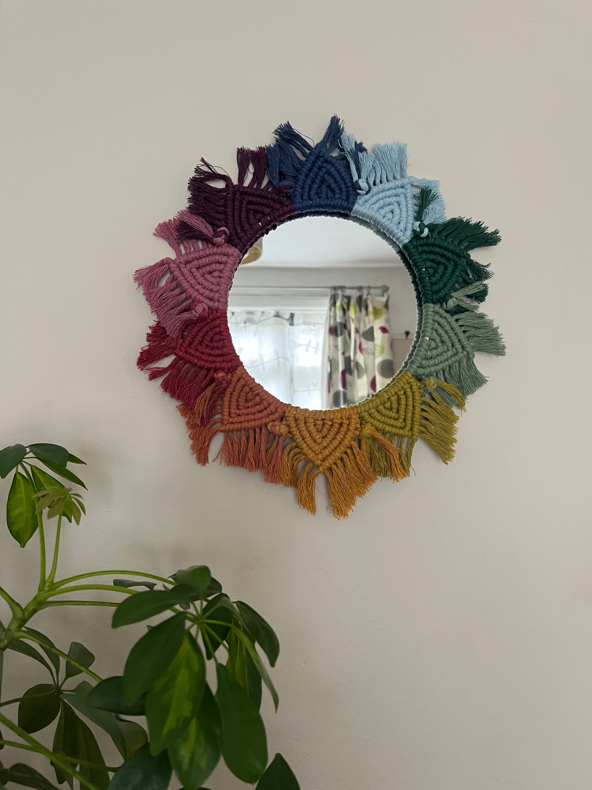 Round wall hanging mirror with rainbow mandala frame, hanging on a beige wall next to a green houseplant.