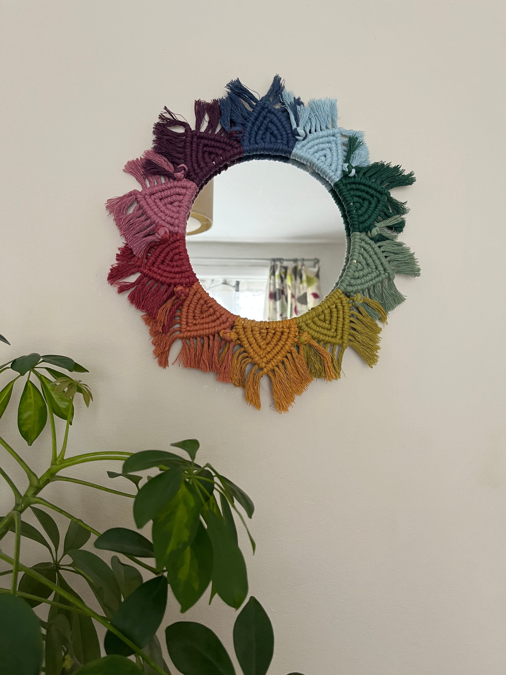 Round wall hanging mirror with rainbow mandala frame, hanging on a beige wall next to a green houseplant.
