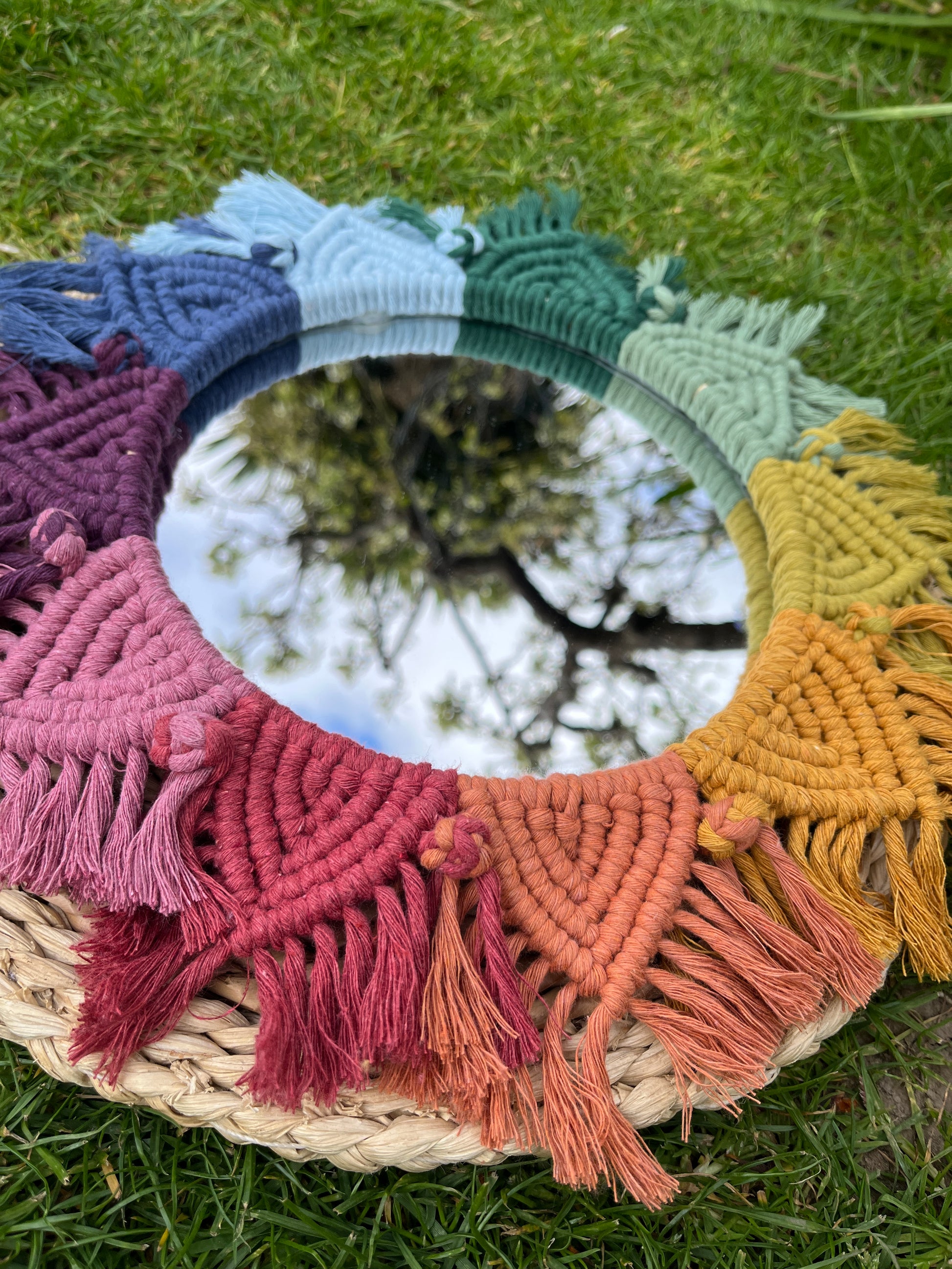 Close up of a round wall hanging mirror with rainbow mandala frame, laying on a wicker placemat outside on the grass. 