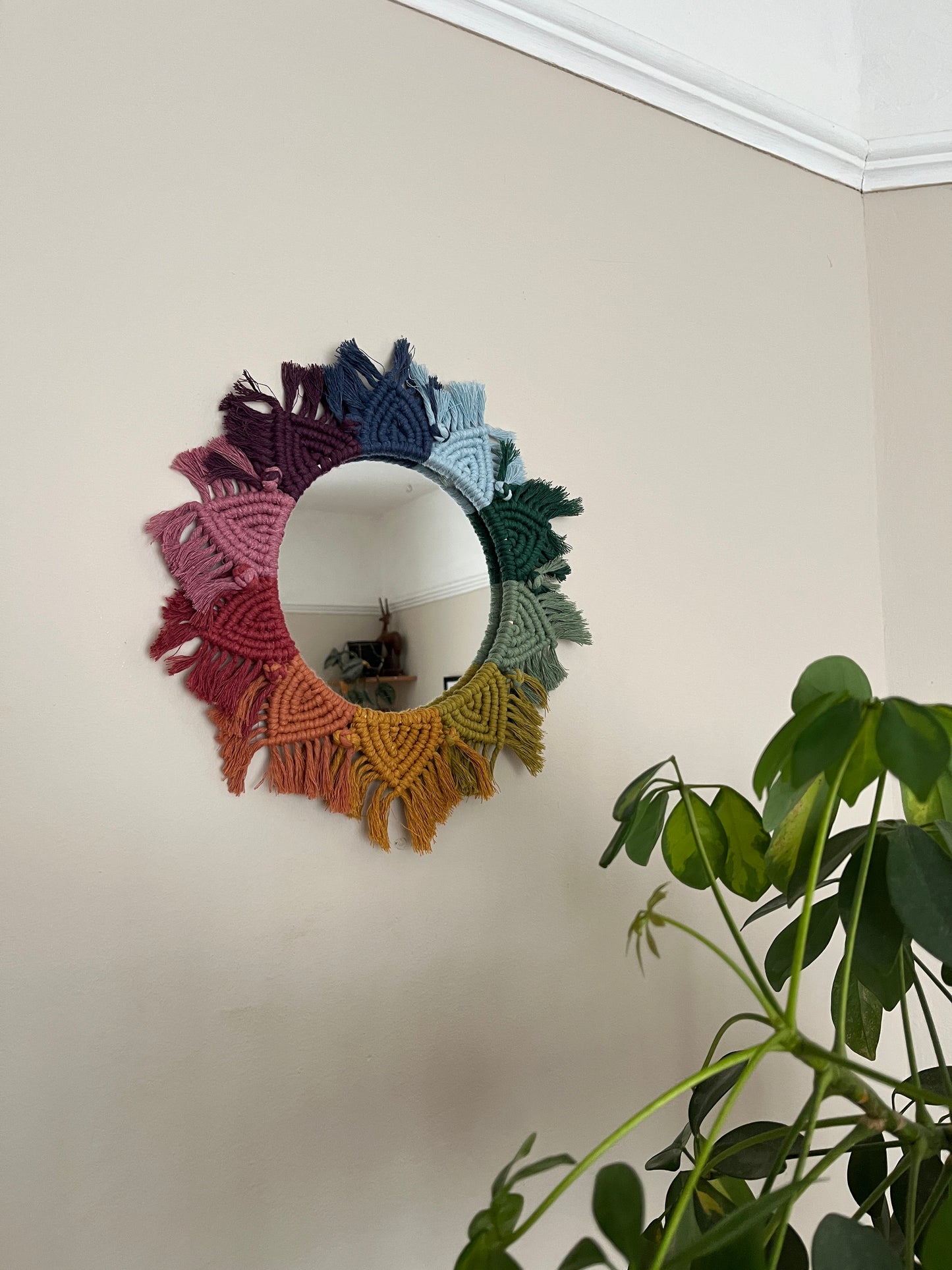Round wall hanging mirror with rainbow mandala frame, hanging on a beige wall next to a green houseplant.