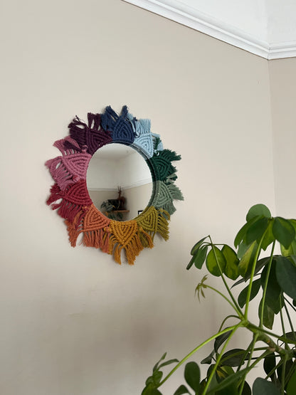 Round wall hanging mirror with rainbow mandala frame, hanging on a beige wall next to a green houseplant.