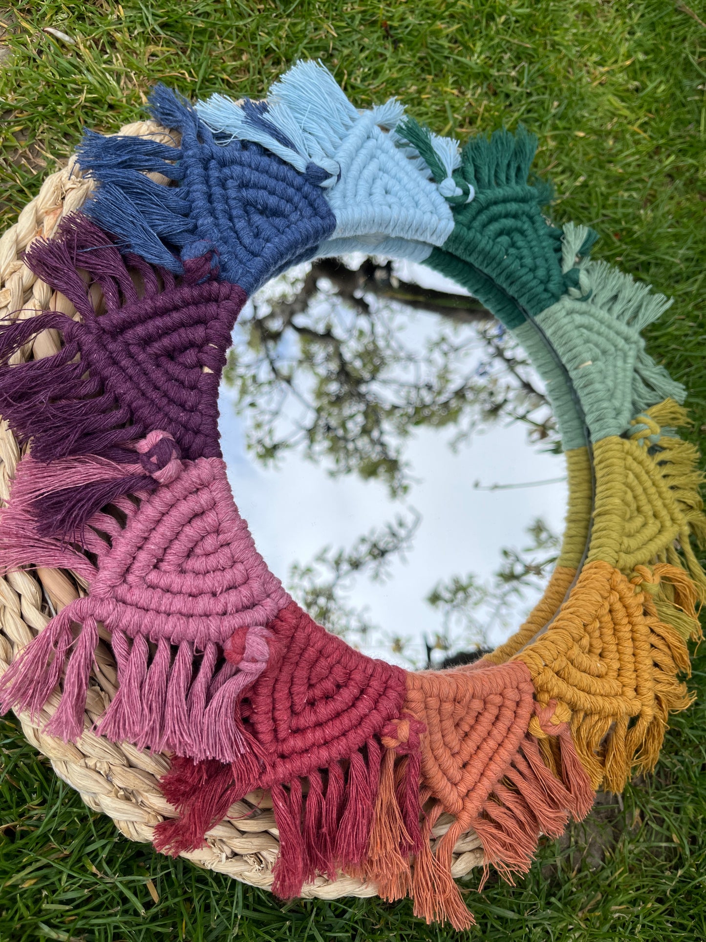 Round wall hanging mirror with rainbow mandala frame, on a round wicker mat outside on green grass. A tree is reflected in the mirror. 