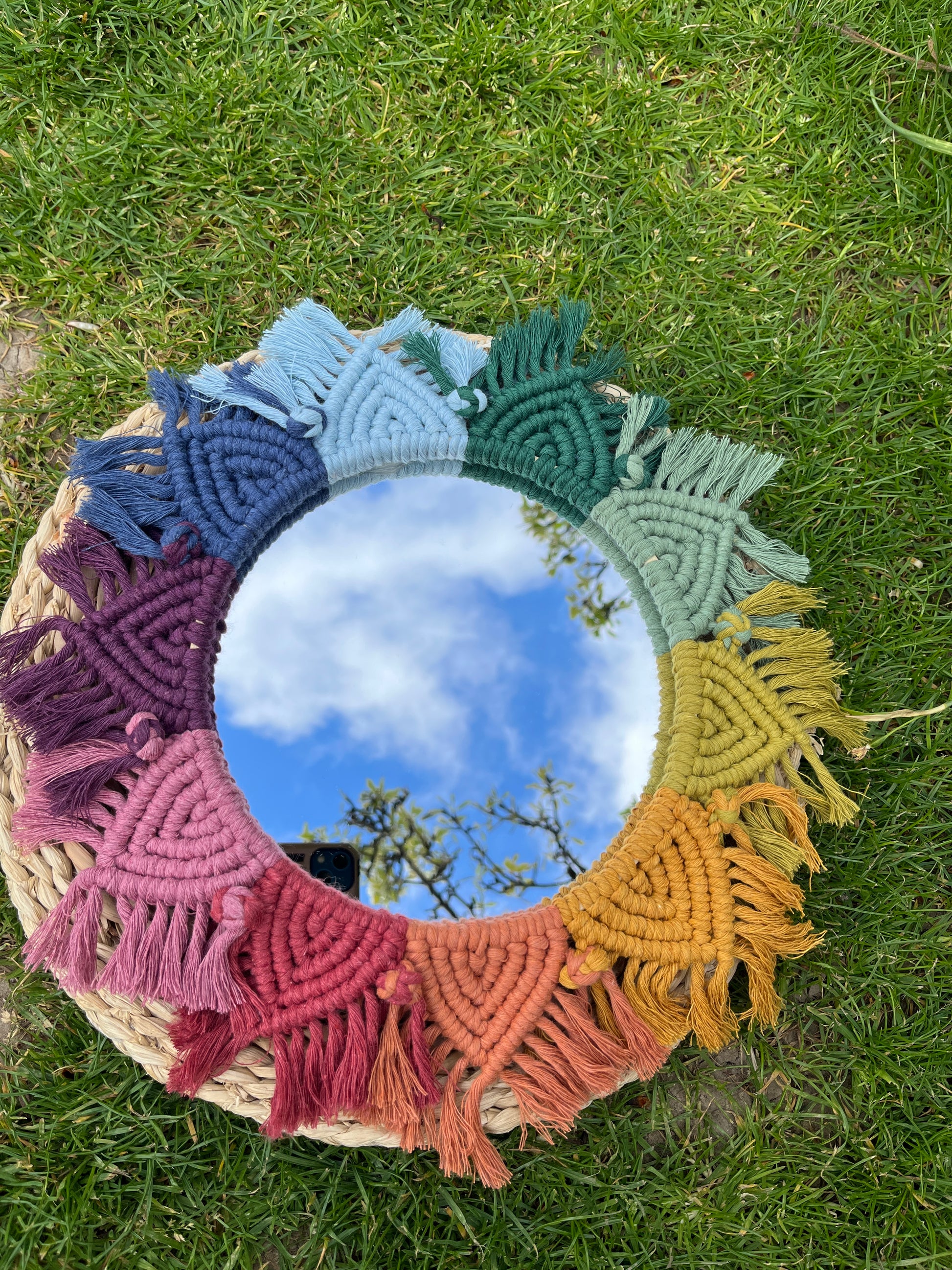 Round wall hanging mirror with rainbow mandala frame, outside in the sunshine on a round wicker surface which is placed on green grass. 