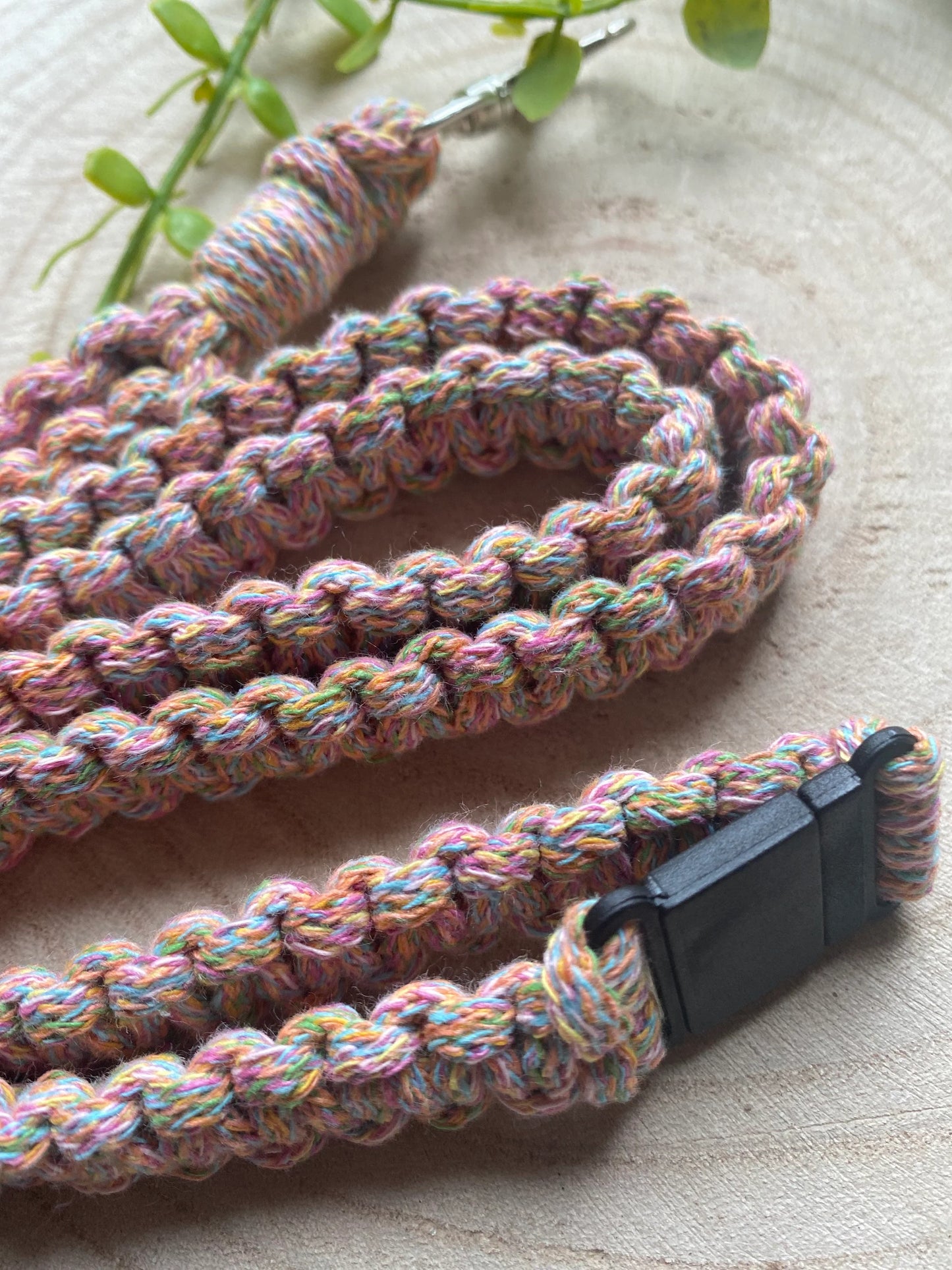 Close-up of a Macra-Made-With-Love Eco friendly lanyard badge holder - 34”, doubling as an eco-friendly ID badge holder with a black plastic clip and metal hook, resting on a light wooden surface. The threads feature a mix of pastel colors. A green plant is visible in the background.