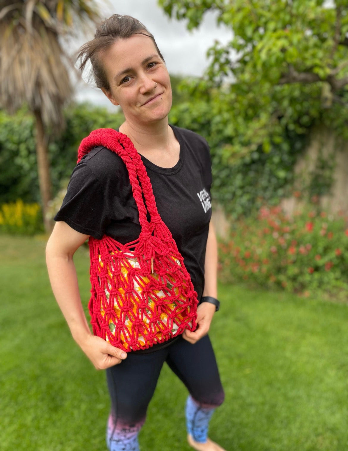 Woman holding a red macrame market bag outdoors with greenery in the background