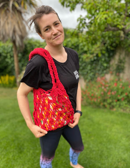 Woman holding a red macrame market bag outdoors with greenery in the background