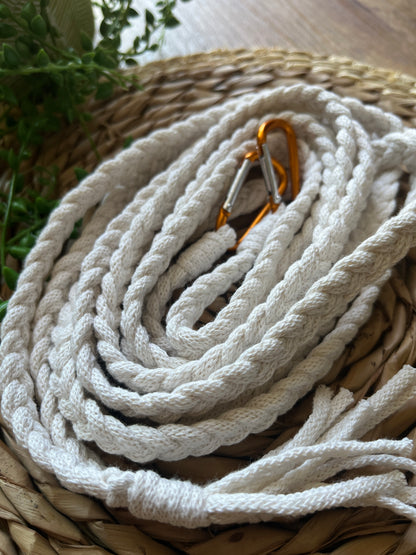 Close up of white braided macrame apron straps with orange carabiner clips, coiled neatly on top of a wicker placemat. 