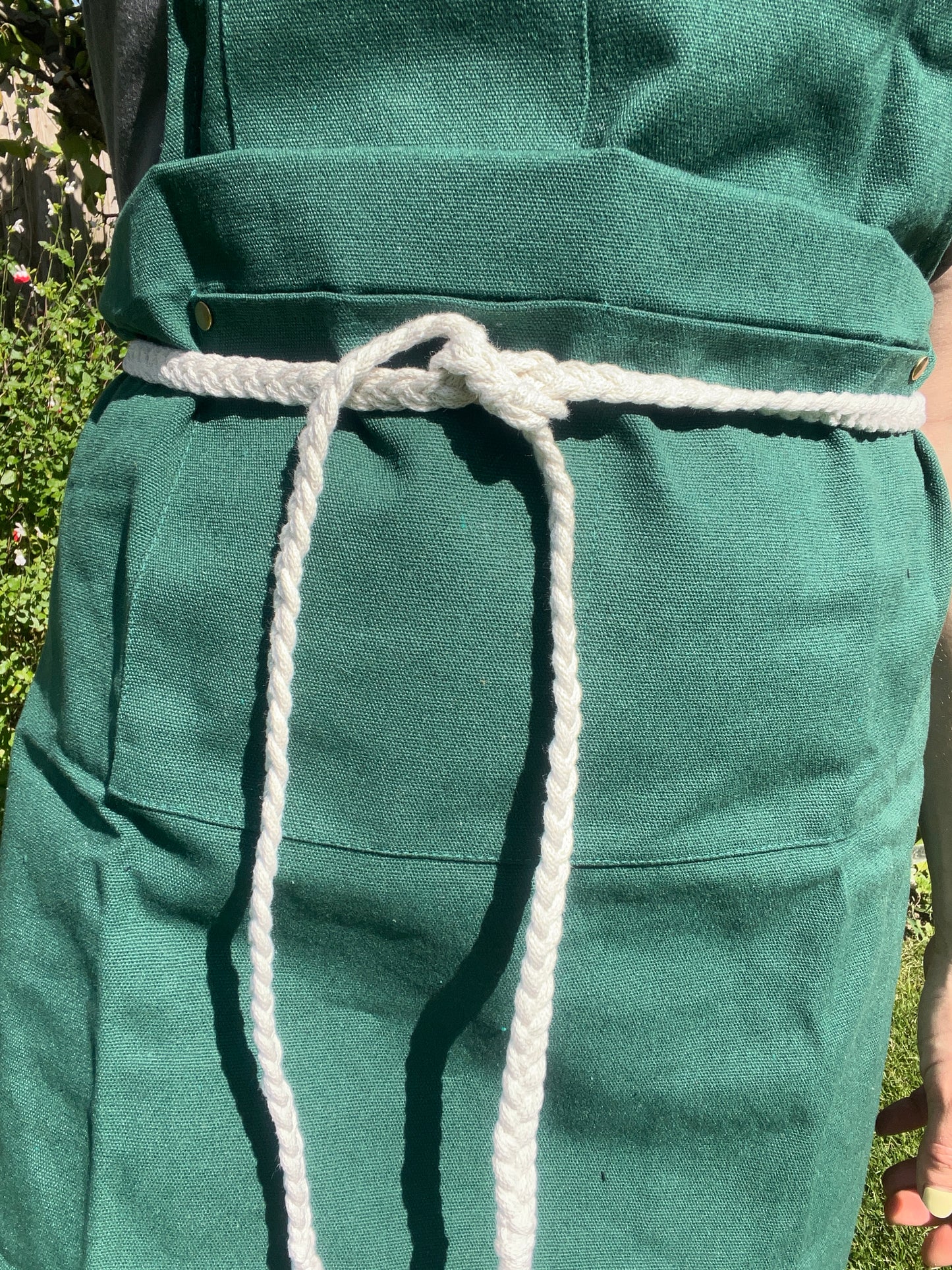 A person stands outside wearing a green apron with a white braided rope waist tie. 