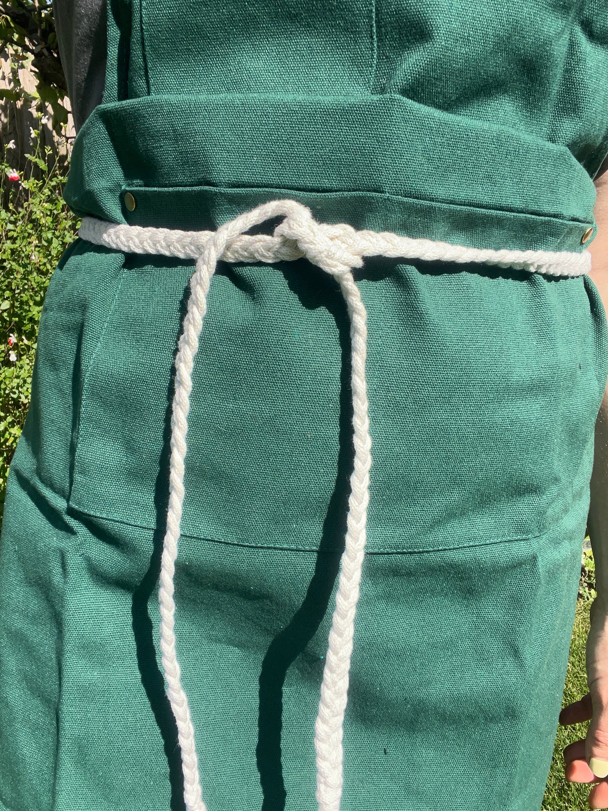 A person stands outside wearing a green apron with a white braided rope waist tie. 