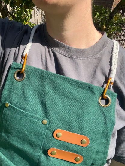 Close up of a person standing outside wearing a green apron with white braided straps attached to the apron with carabiner clips. 