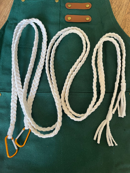 White braided apron straps with orange carabiner clips, coiled on top of a a green apron, which is laid out on the floor. 