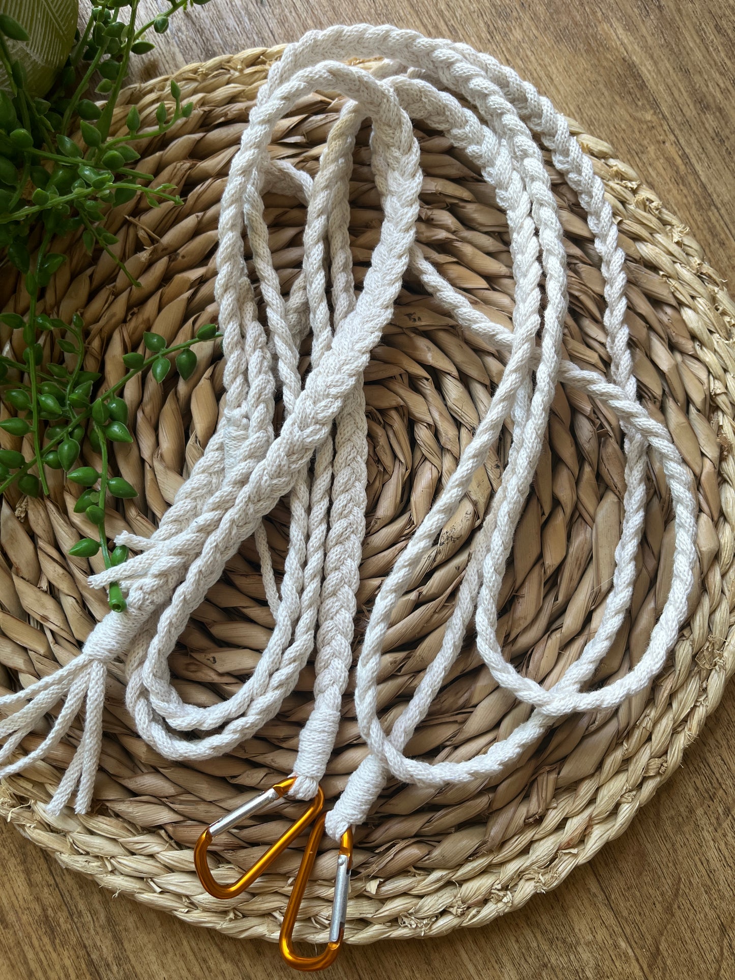 White braided apron straps with orange carabiner clips, coiled on top of a a round wicker mat. 